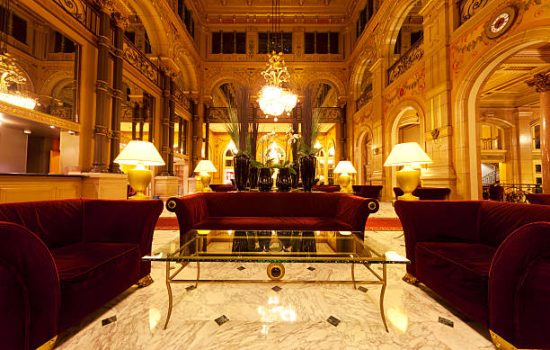 An elegant hotel lobby.  A marbled floor is in the foreground, leading to the furniture grouping of a low glass table and three brown plush sofa couches.  Table lamps and a chandelier provide symmetrical indoor light.  Functional alcoves frame the sides of the image.